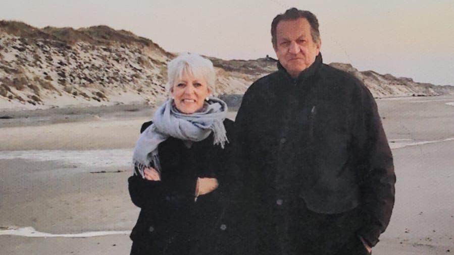 The couple pose together on a beach. It looks like the winter, with them both wearing big coats.