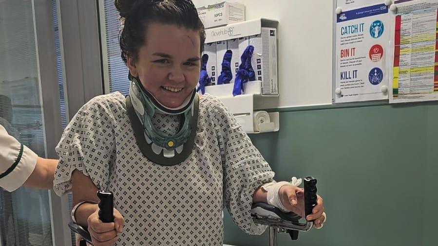 Ms O'Malley using a metal frame to help her walk. She is in hospital, wearing a medical gown and neck brace. She is smiling at the camera.