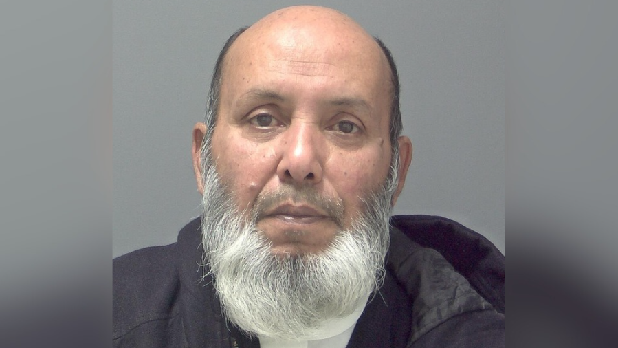 A police custody image of Hafez Ashraf Uddin. He has a grey/white beard, short greying hair on the sides of his head and he is in front of a grey wall. 