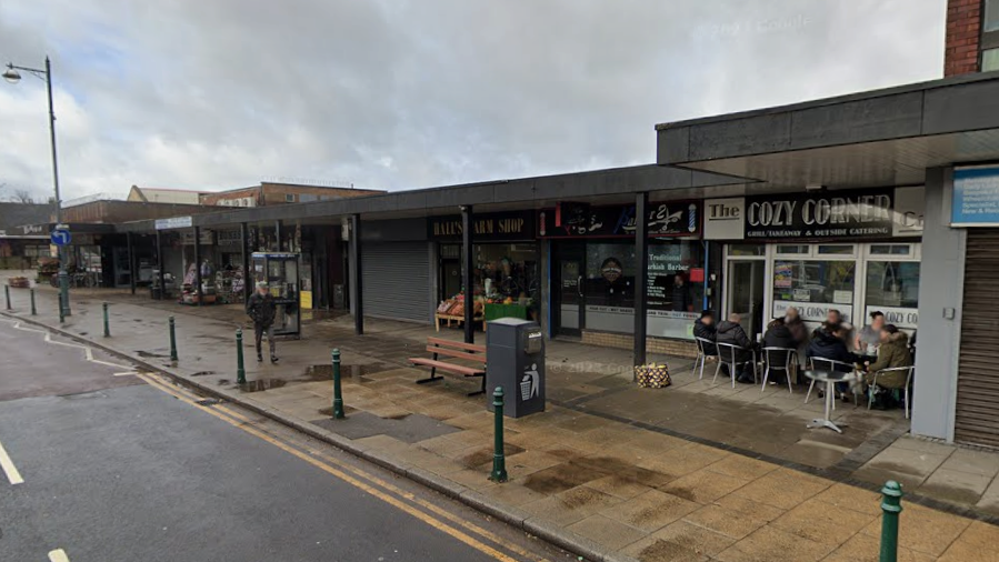 A street scene with single-floor shops to the right hand side. Some people are sitting on chairs outside a cafe, but otherwise, the area looks quite empty.