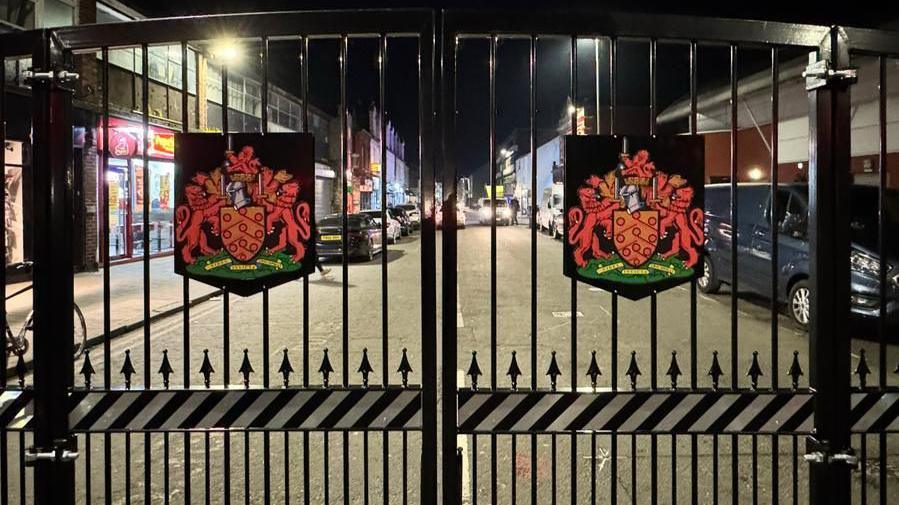 A large black bi-fold set of gates with coats of arms on each one on a commercial street in Gloucester city centre. Parked cars can be seen on the road, which has fast food venues on.