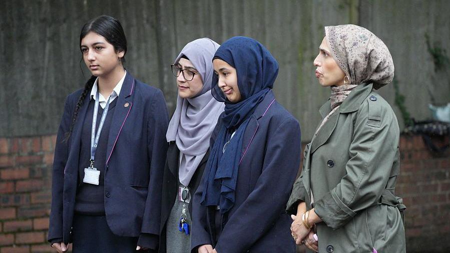 Three females in Muslim hijab headscarves and another Asian girl stand in silence and sombrely at an event at the scene of the attack