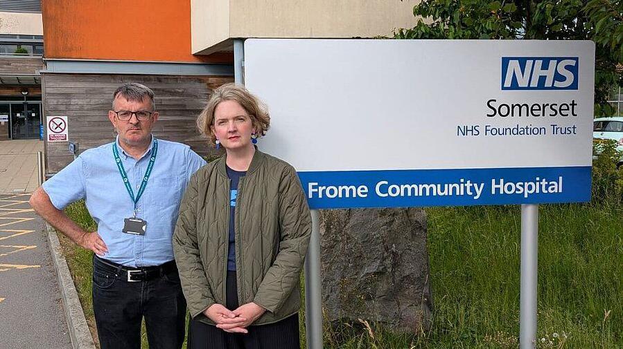 A man and a woman standing side by side. The man has his hands on his hips and the woman has her hands in front of her. They are standing in front of the sign for Frome Community Hospital.