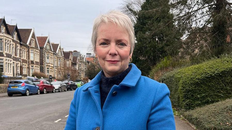 Karin Smyth standing on the pavement on a residential street. She is wearing a dark blue turtleneck jumper underneath a cobalt blue button up coat. She has short white hair, is wearing small hoop earrings, and smiling at the camera.