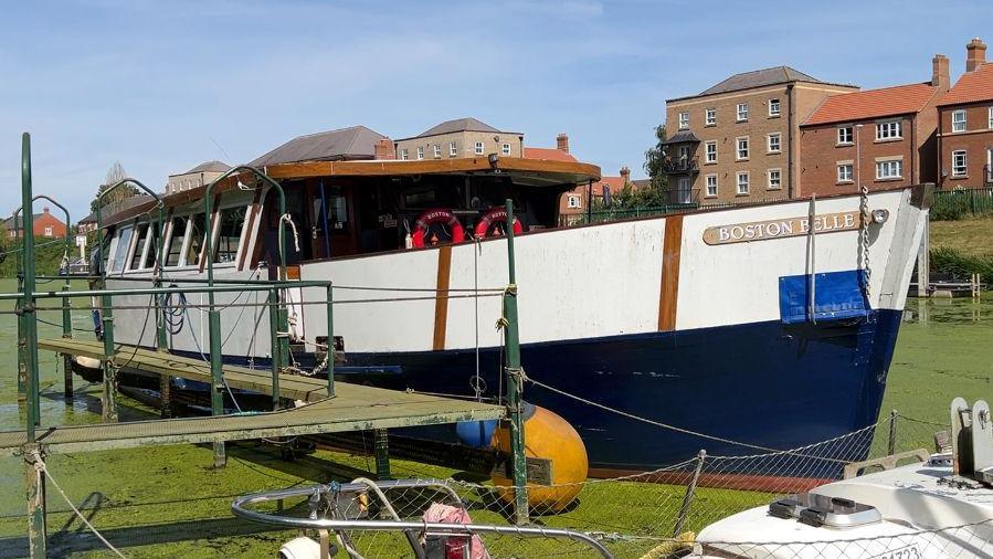 The Boston Belle amid a carpet of green weeds on the River Witham. The boat has a blue and white Hull and a wooden cabin with widows down the side. A wooden gang plank leads to the bank