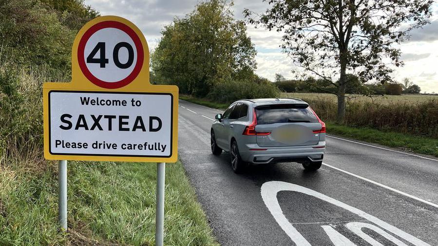 A Welcome to Saxtead sign showing the new 40mph speed limit. A grey car is driving past it as the photo is taken. 