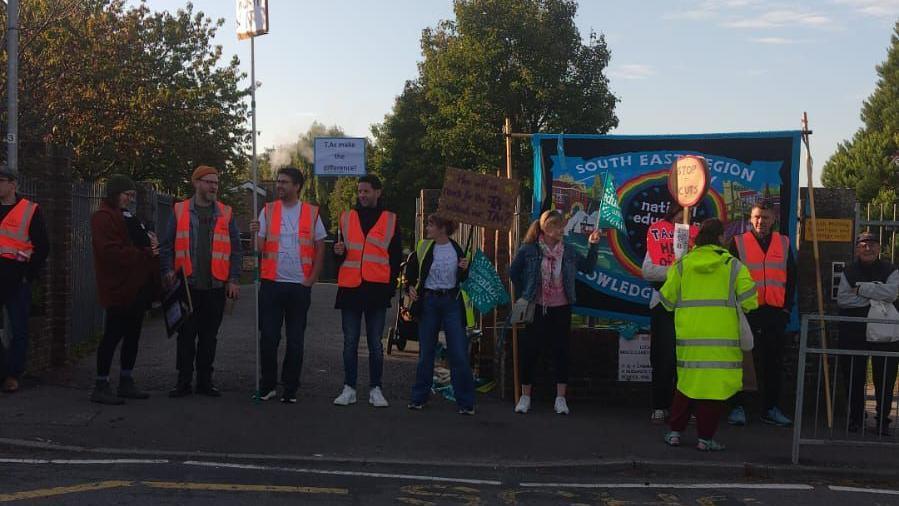 People in high visibility jackets in a line at the side of a road, beside the entrance to South Malling Primary School. Some of them are carrying homemade signs.