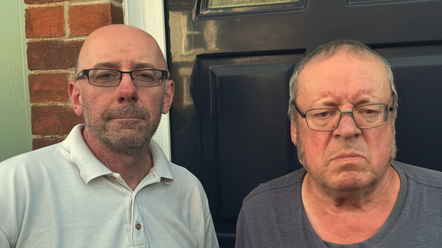 A close-up head and shoulders image of two men. They are both wearing glasses and standing in front of a blue front door. They are looking into the camera.