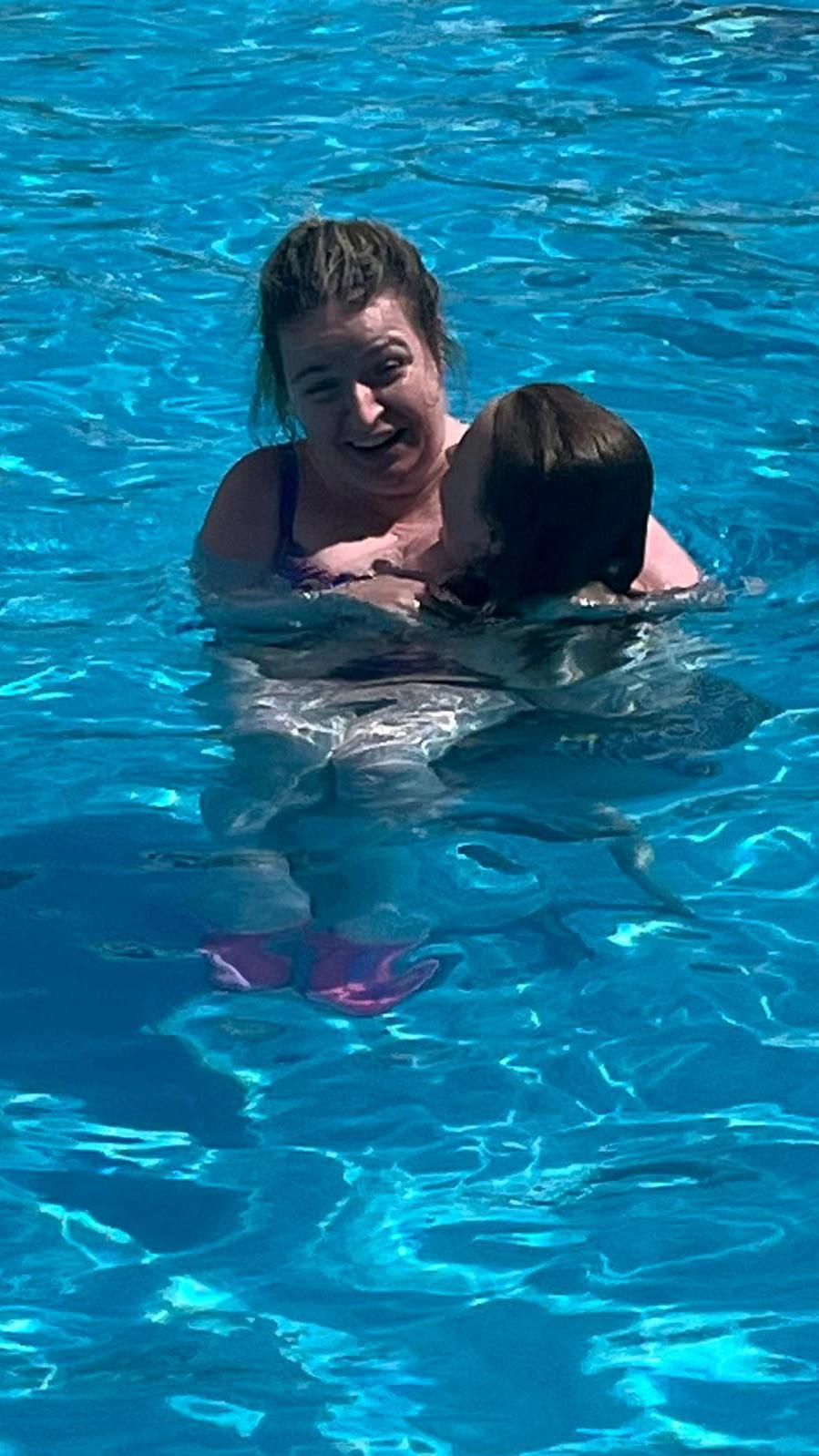 A woman in her forties with dark blonde hair which is pulled back, stands in a blue shimmering swimming pool in the sunshine with the water almost up to her shoulders. She is smiling and her eyes are closed. She is holding on to a teenage girl who has long brown hair, which is wet, and who is facing mostly towards the woman. The girl is in the water up to her neck and both of their bodies and limbs are not clearly visible, as they are distorted by the water.