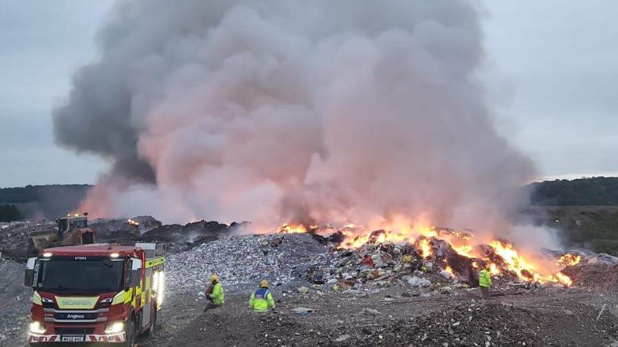 Fire engine and firefighters near pile of rubbish that is on fire