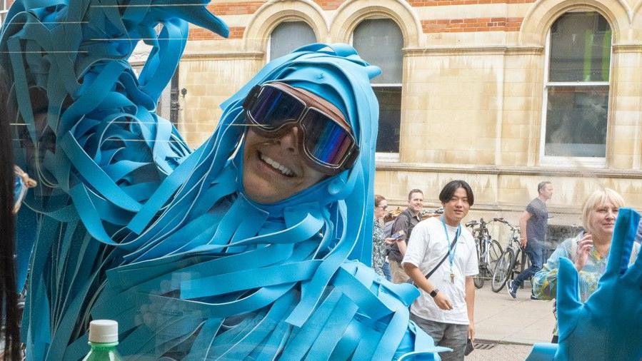 A man is dressed in an elaborate costume made of long strips of blue fabric ans is wearing large goggles. He is waving through a window at someone sitting inside. There are people walking on the street behind him