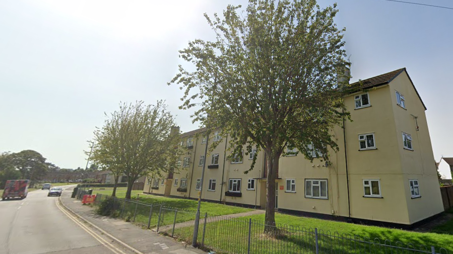 A Google Maps screenshot of the outside of a block of flats. The building is three storeys tall, is quite wide and light yellow in colour. There are three trees in front of the building, and the picture has been taken on a sunny day. 