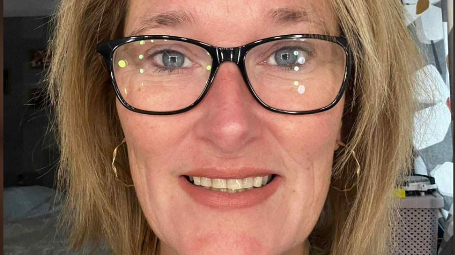 A fair-haired woman with glasses and hooped gold earrings smiles directly at the camera.