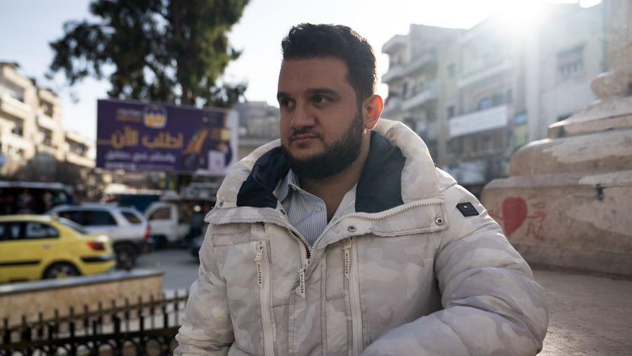 Young bearded man in white coat stands next to a road in a built-up part of Idlib