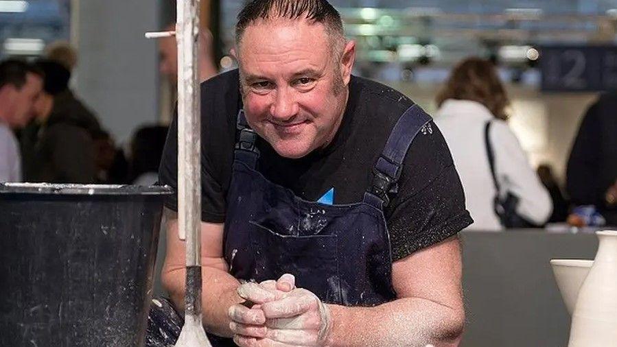 Keith Brymer Jones, wearing a black t-shirt and dark-coloured apron, can be seen with his hands clasped around a piece of clay. There are pots next to him, and a plastic bucket, and people in the background.