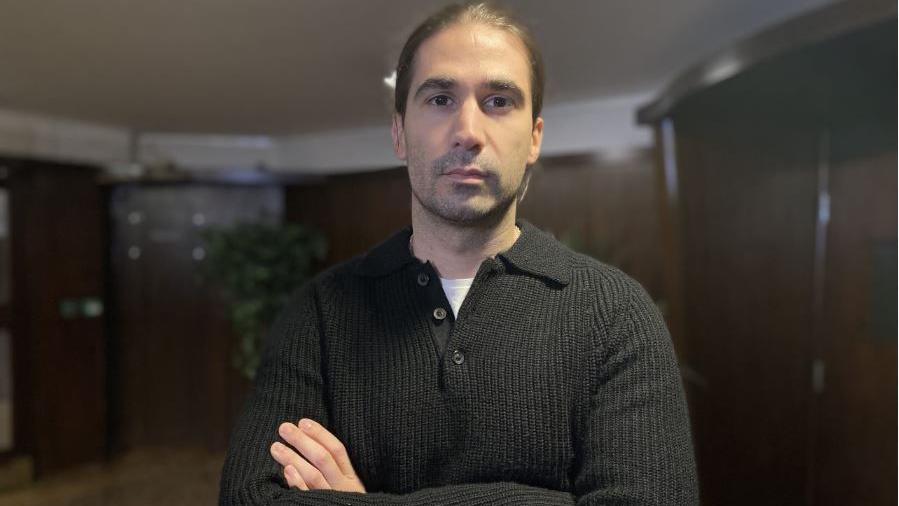 Abrahan Marquez looking into the camera with his arms folded, wearing a black jumper, in a communal area of his flats