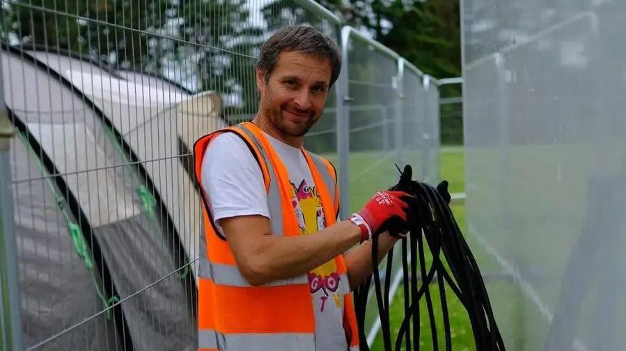 Stu Housley, wears a high-vis and gloves, white t-shirt, he holds black ropes in his left hand and smiles at the camera.