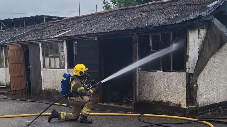 A firefighter in full gear shown hosing down the inside of a low, burned-out structure which appears to be a workshop. 