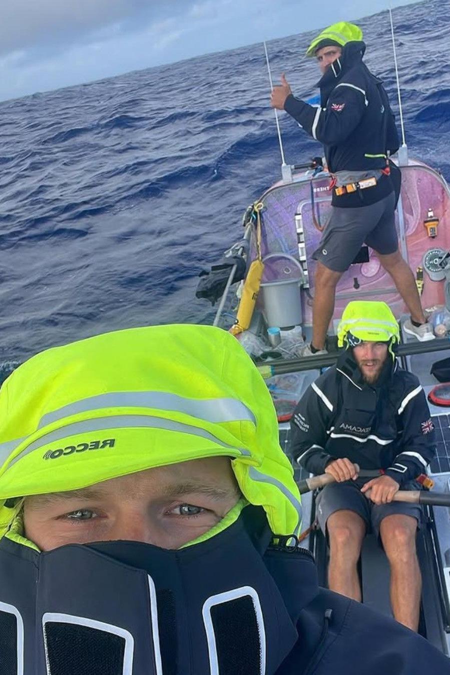 The three brothers are in the boat in rough sea conditions. They have shorts on but are wearing jackets with the hoods up.