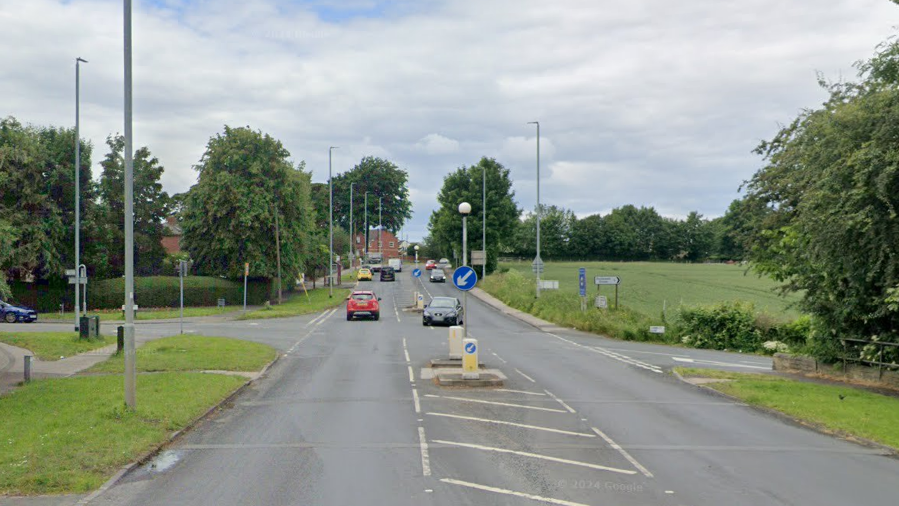 A61 junction with Jumbles Lane in Lofthouse, with the road fairly busy with vehicles. A black car is turning right, with a red car continuing along the A61.  There's grass and trees on both sides of the road. 