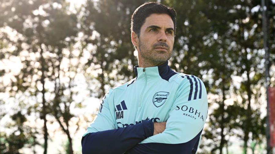 Mikel Arteta stood with his arms folded looked away from the camera against a backdrop of trees wearing a light blue arsenal training top.