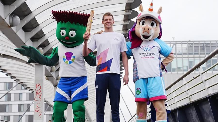 Donnchadh Scott na sheasamh air drochaid còmhla ri mascot Geamaichean a' Cho-fhlaitheis.