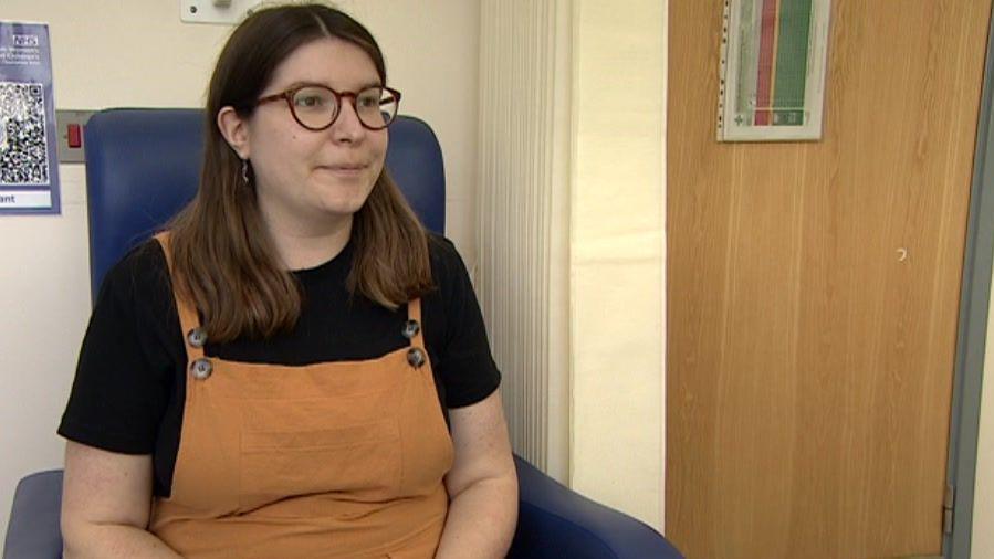 Chloe Le Breton looking at a medic who is out of view. She is sat down and is wearing brown dungarees and a black t-shirt and had a brown door behind her. 