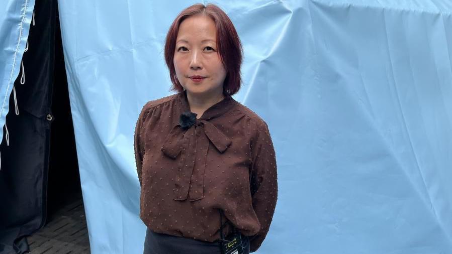 A Japanese woman stands in front of a light blue fabric backdrop with hands behind their back. She is wearing a brown long-sleeved blouse with a bow-tie neckline and dark trousers