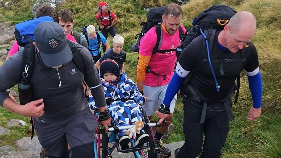 Eight people going up Ben Nevis, in climbing gear with equipment, carrying Ted in his wheelchair, he is wrapped in blue, white and white clothes.
