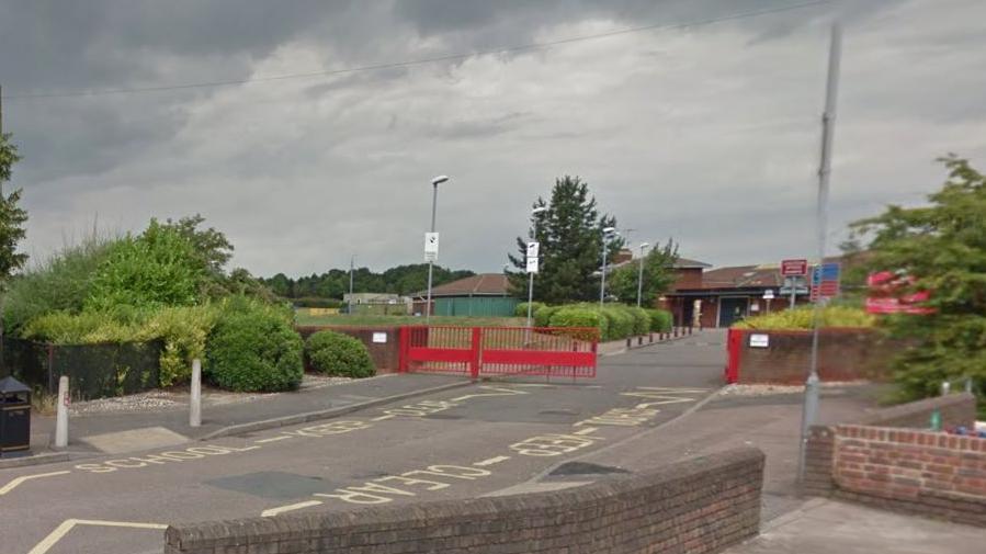 Red metal railings across a road, with part of a gate open in front of a low-level red brick building with bushes and grass outside.