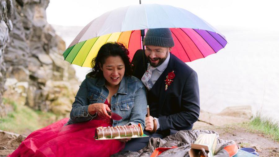 A man and a woman sit together on the floor next to a large rock. The man is wearing a suit and holding a rainbow umbrella and the woman is wearing a formal red dress and denim jacket and is slicing into a chocolate cake