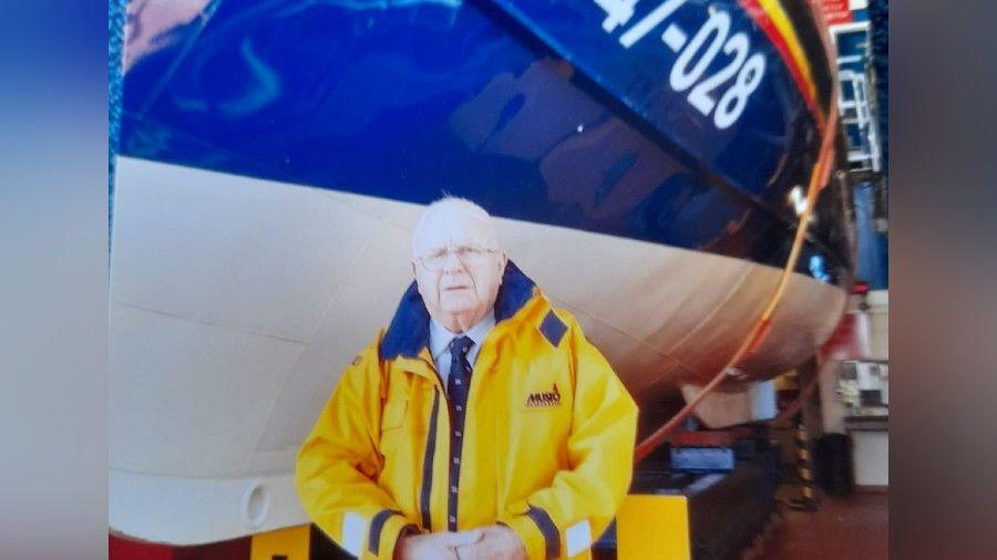 Bob McLaughlin standing in front of an RNLI boat, which has been hoisted out of the water into the lifeboat station. He has white short hair and is wearing glasses and a yellow waterproof jacket over a shirt and blue tie.