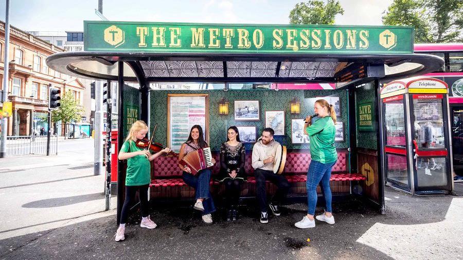 Musicians sat on a bus stop, which has been decorated to appear as a bar. The sign reads "The Metro Sessions".