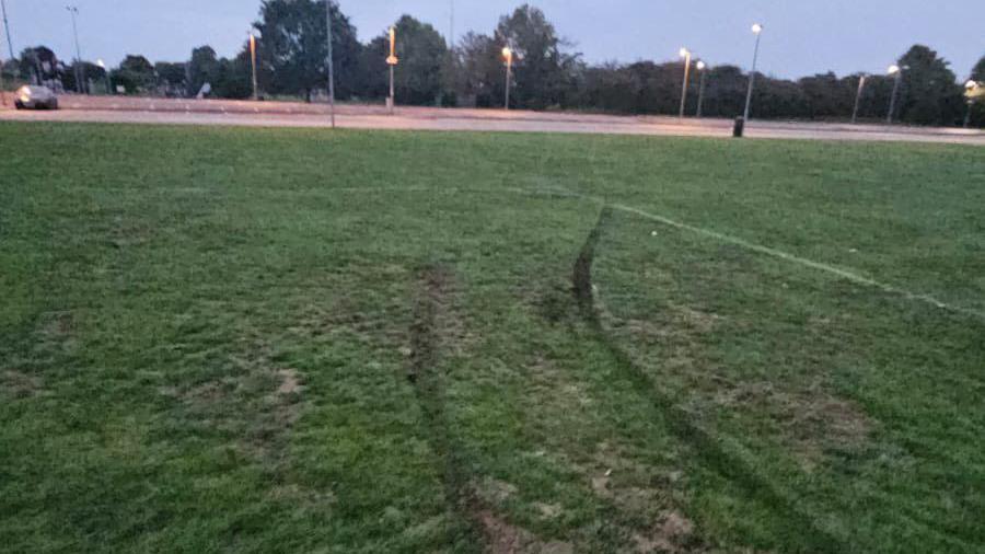 A grassy football pitch that has been visibly damaged by a vehicle. Two deep, parallel tire tracks cut across the field, disturbing the soil and flattening the grass. In the background, there’s a car park with several light poles and trees, and the sky appears dim and the streetlights are on. The scene shows the kind of vandalism Littleport Rangers FC have been dealing with, a vehicle driving onto the pitch and tearing up the surface, making it unsafe or unusable for play.