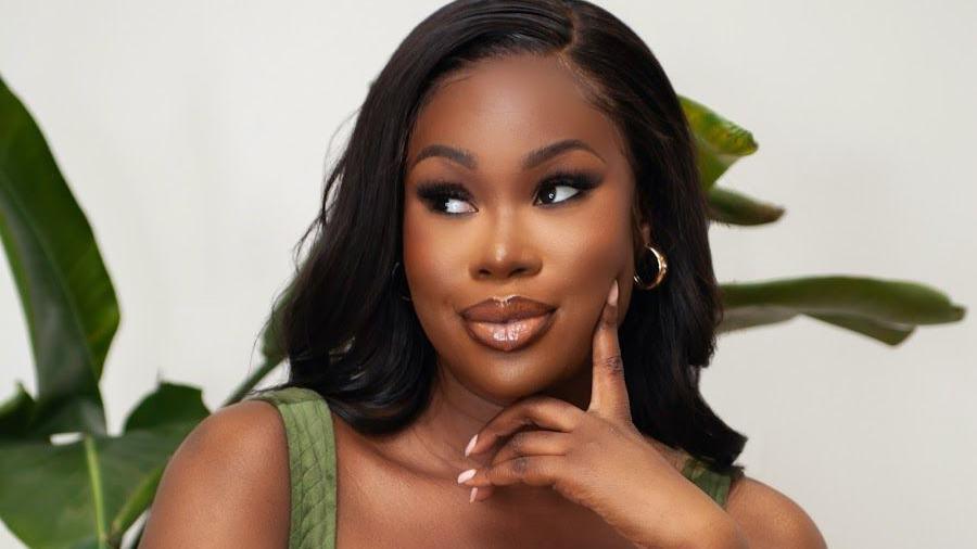 Dami Olonisakin, poses with her hand framing her face in a professionally shot photograph. She has curled dark hair, dark eye make-up and shiny lip gloss on, with a green sleeveless dress.