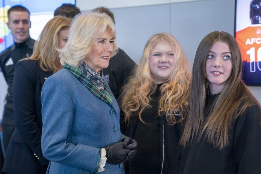 Queen Camilla, wearing a light blue jacket and a flash of tartan, smiling, with two young women.