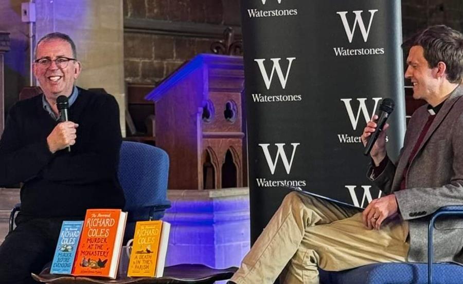 Richard Coles with short dark hair and glasses, smiling while holding a microphone and wearing a black sweater. The Reverend Tom Houston, with short dark hair and a grey jacket, sits opposite. Books by Mr Coles are on display in the foreground, and a Waterstones logo is visible behind