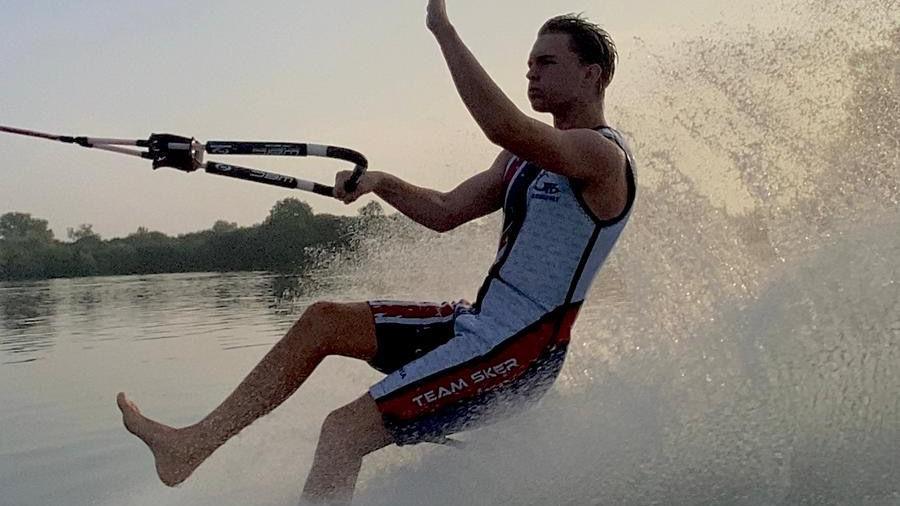 A boy wearing a neoprene suit is skiing on the water with his bare feet. He is holding on to a rope while his left arm is held up. The water is splashing behind him.