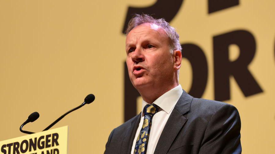 A man wearing a dark suit and white shirt with a blue tie with yellow flowers pictured. He is talking into a microphone with a yellow screen in the background.