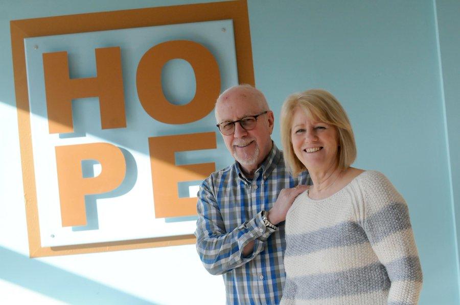 Orange sign reads 'HOPE' on a blue wall. To the right, a white couple stand - the man has his hand on the woman's shoulder.