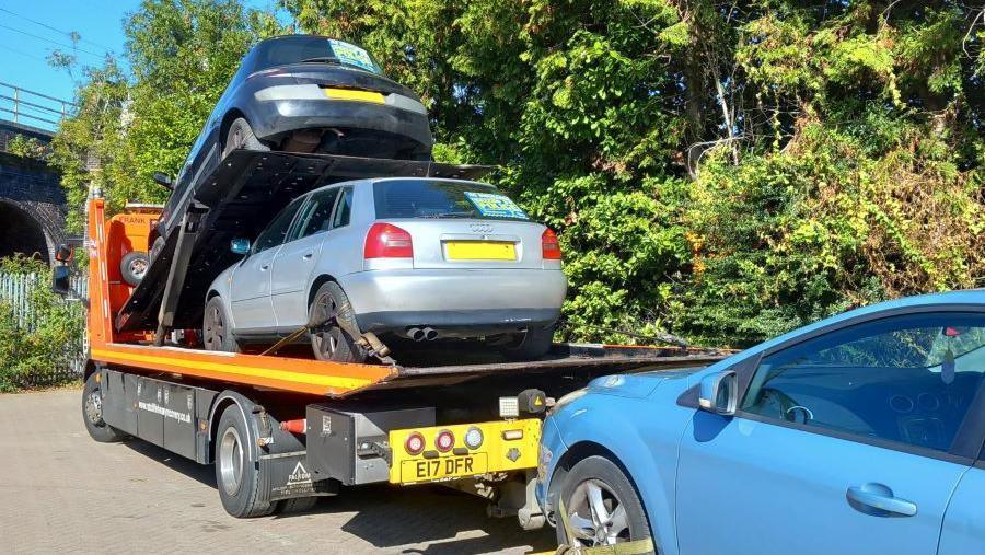 Two cars, one black and one grey, are on the back of a lorry which is orange and black. A blue car is on the left next to rows of trees.
