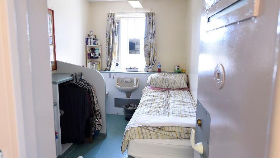 Open cell in Hydebank. There is a single bed with a duvet on it, a sink, window with curtains and storage area with clothes on hangers. 