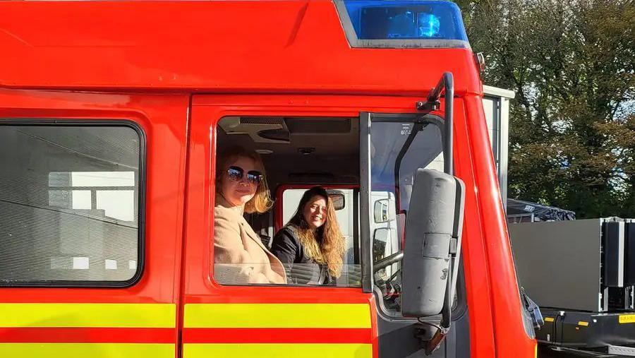 Two brown-haired women sitting inside a red fire engine with yellow emergency service stripes. The one behind the wheel is wearing sunglasses. 