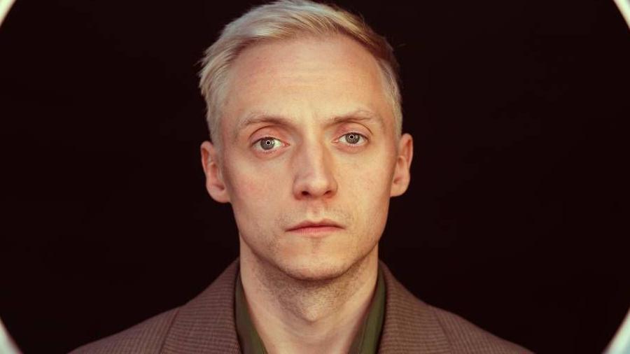 A portrait of a man with short, light blonde hair. He's wearing a brown textured jacket over a dark green collared shirt.
