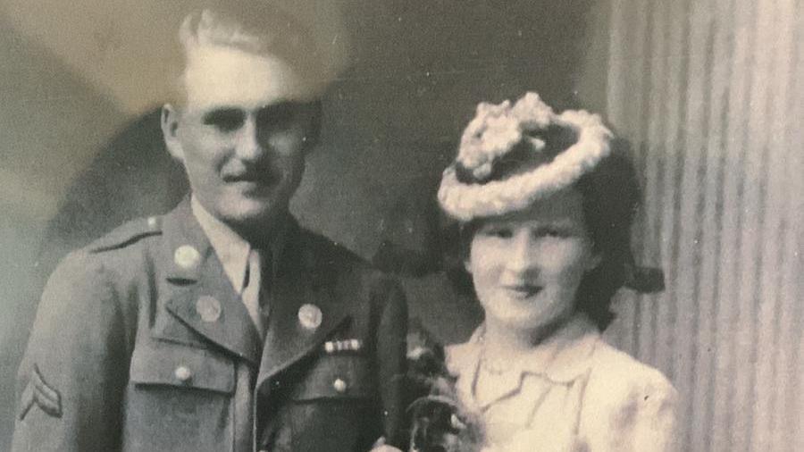 A black and white photograph of a couple. Emlyn is on the left, wearing a hat, with Chester in uniform on the left. They are both smiling at the camera and she is holding on to his arm.