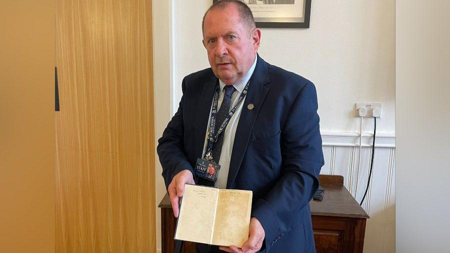 Headmaster Andrew Keep holding the book open on the first page with the inscription. Mr Keen is wearing a blue jacket, tie and a white shirt. He has dark short hair and a moustache.