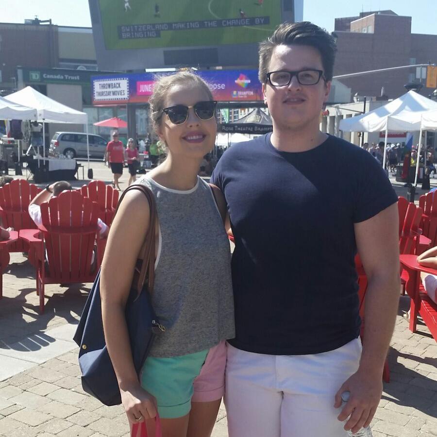 Two people smile as they look into the camera in front of an outdoor seating area