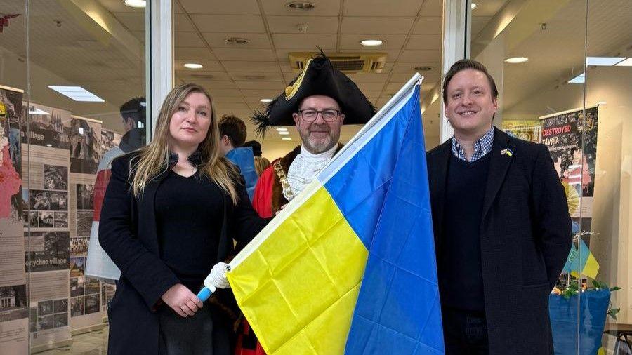 Antonina Grebeniuk standing beside the Lord Mayor of Bristol, who is wearing his red mayoral robes, a gold chain and a black hat. They are holding a Ukrainian flag and standing in front of the door to the exhibition.