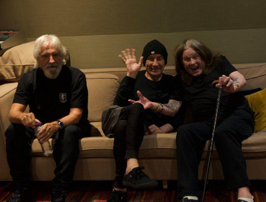 Three men sit on a beige sofa. They are all wearing all-black outfits. The man on the left has grey hair and a grey beard. The man in the centre has a black beanie hat. The man on the right has shoulder-length greying hair and a walking stick.