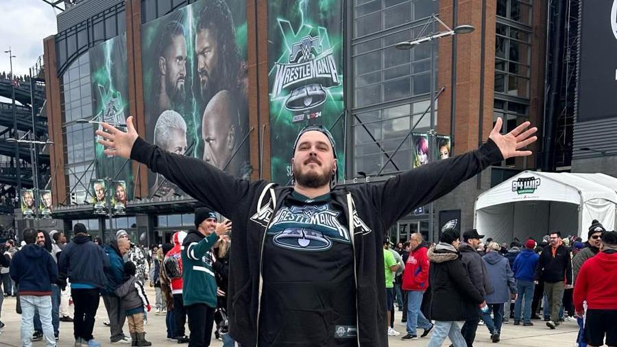 A picture of Jonathan Elgie in a Wrestlemania top with arms spread out in front of the Wrestlemania sign